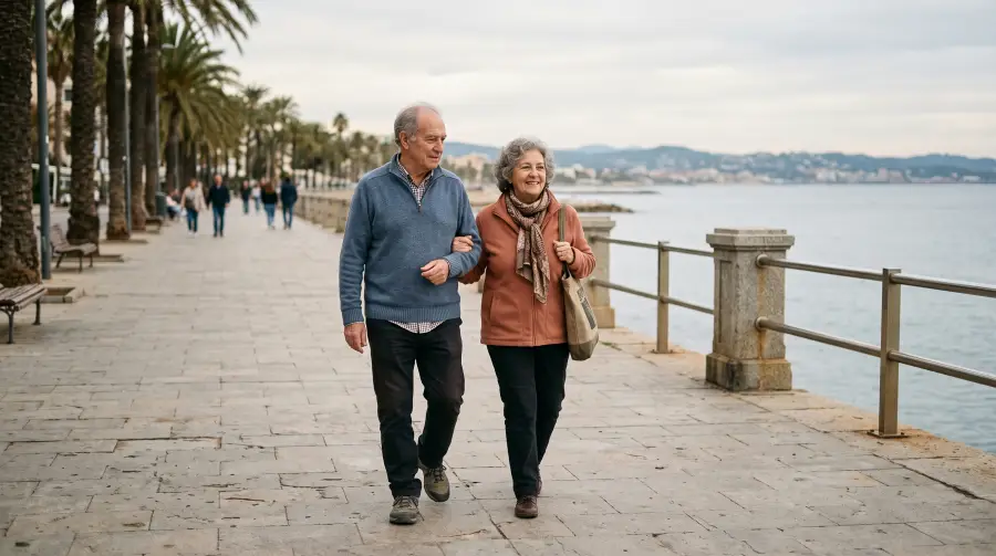 Una pareja de jubilados paseando del brazo por un paseo marítimo en otoño.